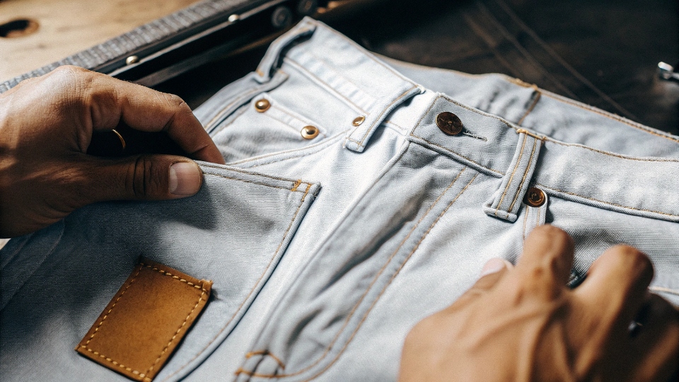 A Guide to Identifying High-Quality Denim Jeans A close-up of a person's hands inspecting the key quality markers of a jean: the metal rivet, the thick fabric, and the leather patch.