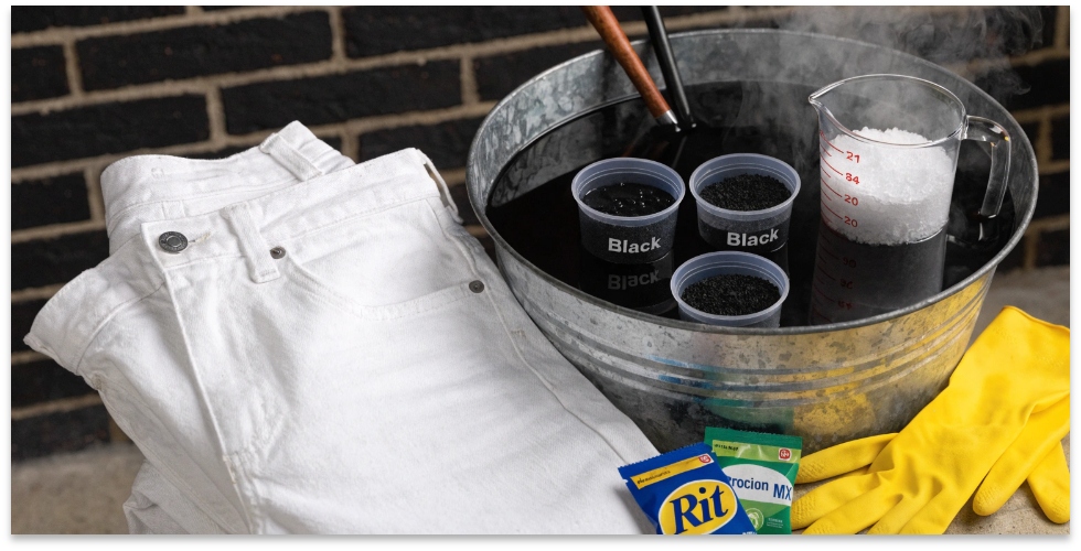 Supplies for dyeing white jeans black A pair of white jeans next to a bucket of black dye and rubber gloves